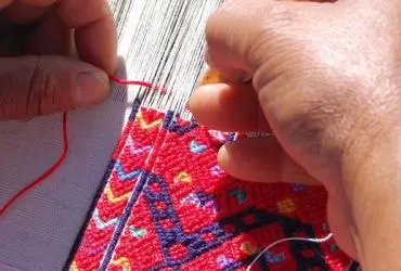 Person's hands weaving a piece of fabric on a table, with threads of various colors
