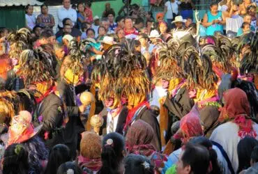 Large crowd of people wearing colorful costumes and hats, gathered around a building with a green wall in the background