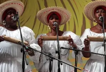 Three women in white dresses and straw hats singing into microphones, each holding a musical instrument