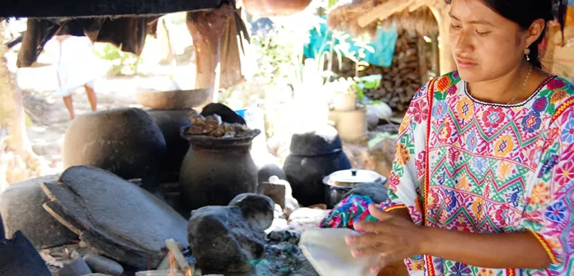 Woman in a colorful dress standing in front of a stove, surrounded by stones, pots, and other objects