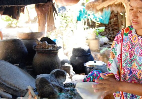 Woman in a colorful dress standing in front of a stove, surrounded by stones, pots, and other objects